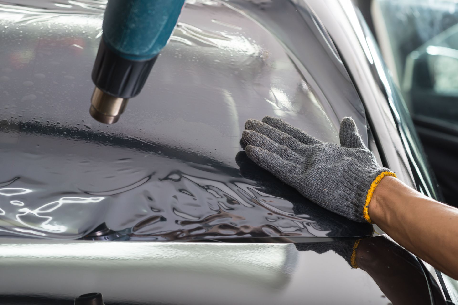 A gloved hand smooths window tint film onto a car windshield while a heat gun warms the surface.