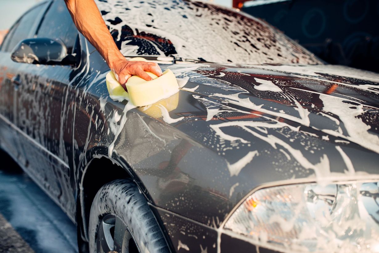 A person using a yellow sponge to wash a soapy, dark-colored car.