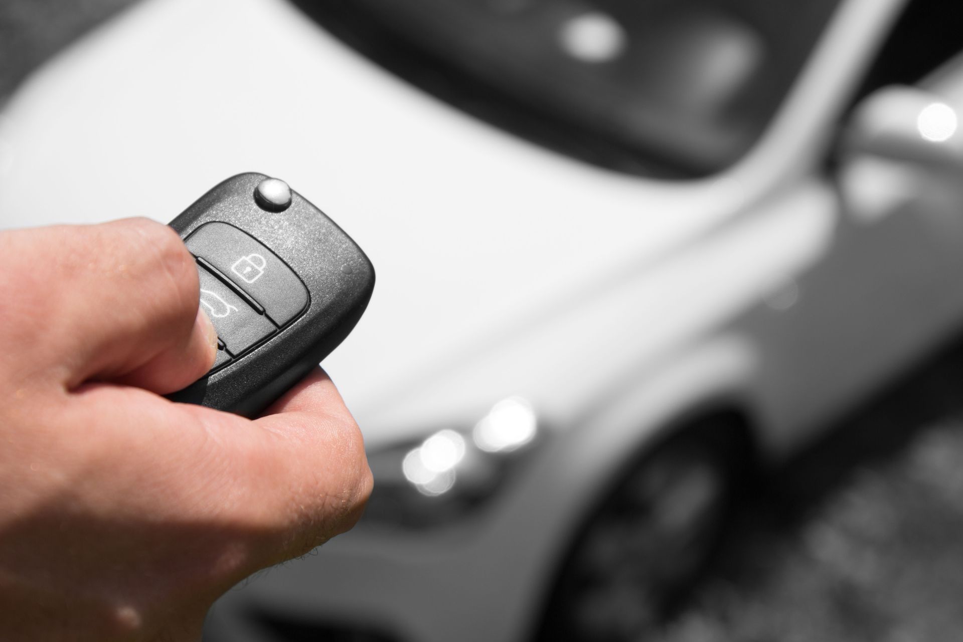 A hand holds a black car key fob, with a blurred white car in the background.