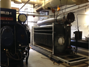 Boiler room with large industrial boiler, piping, and gauges. Dark machinery in a concrete room.