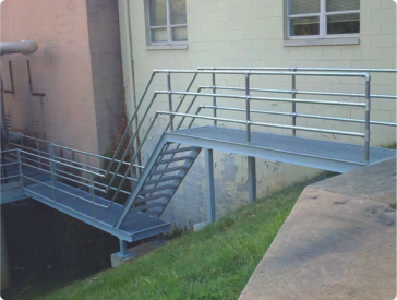 Exterior metal stairs and walkway with handrails next to a building on a grassy slope.