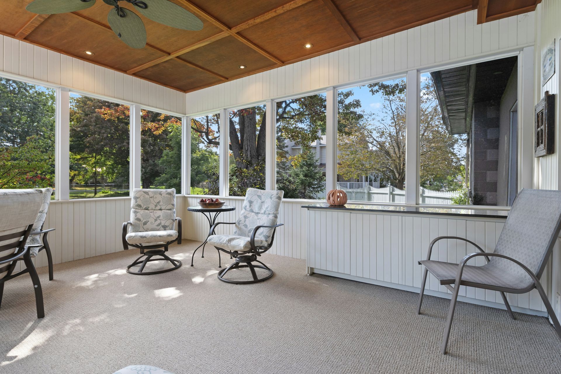 Sunroom with white walls, beige carpet, and outdoor seating, overlooking a green yard.