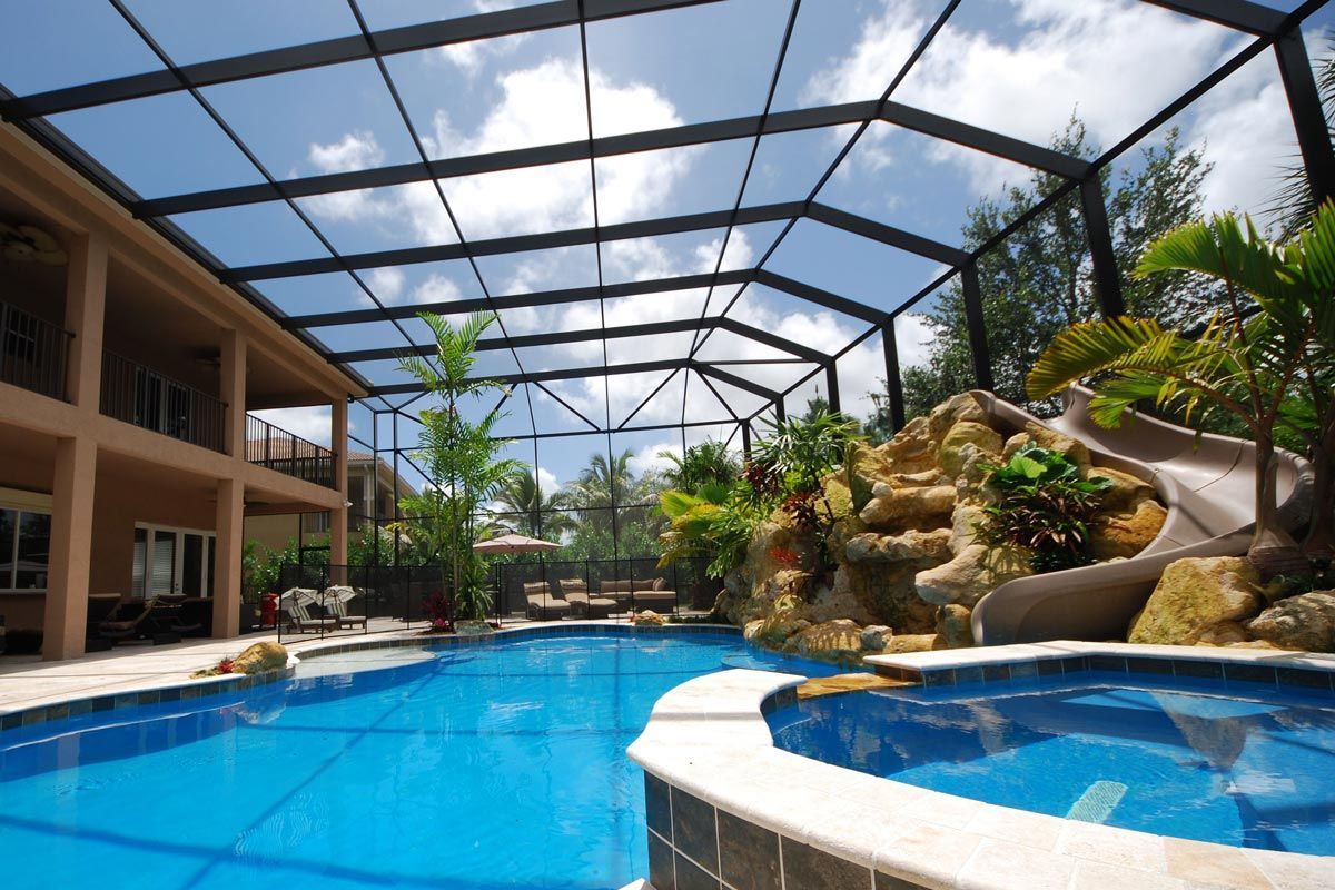 Blue pool and jacuzzi enclosed by a screened structure with a slide and tropical landscaping.