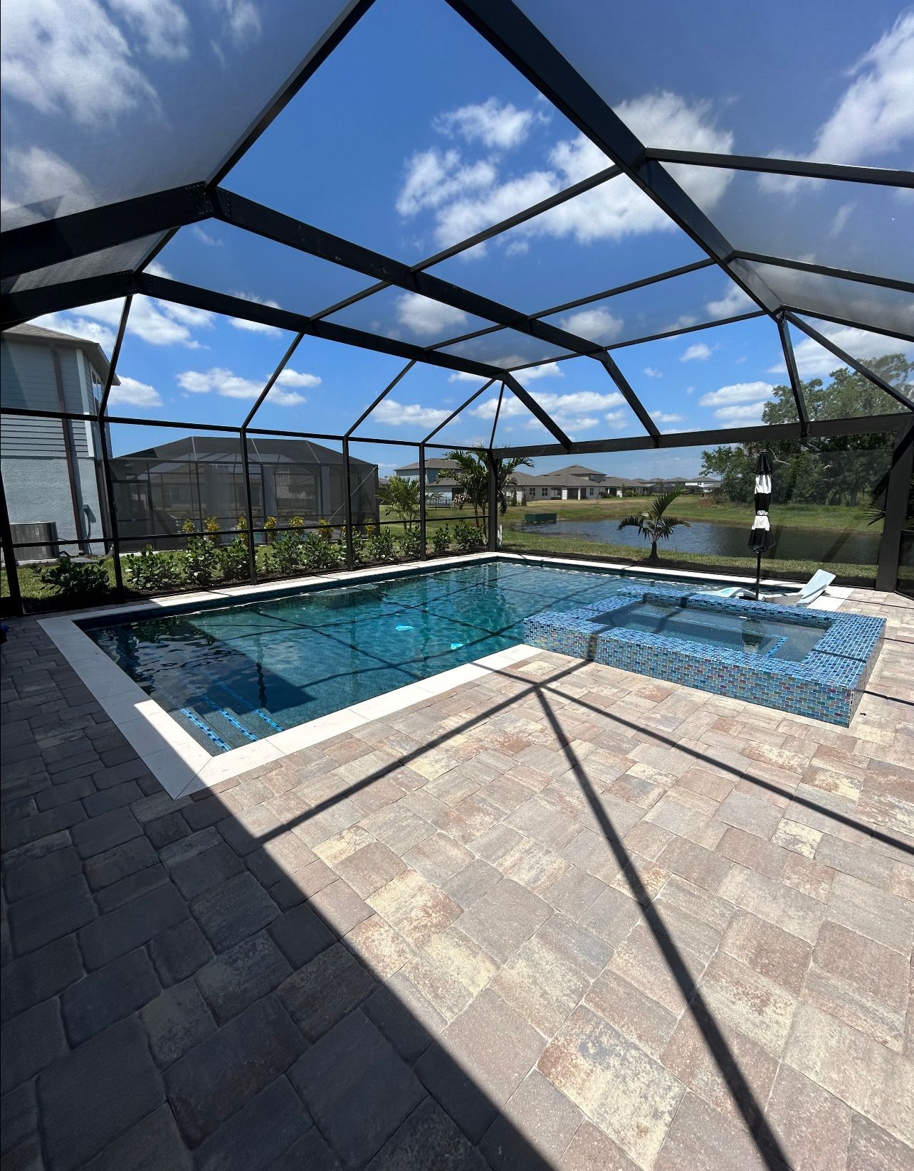 Pool with a jacuzzi enclosed in a screened lanai, surrounded by brick pavers, under a bright blue sky.