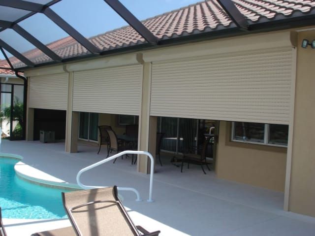 Beige roll-down shutters protect an outdoor patio with a pool, under a glass roof.