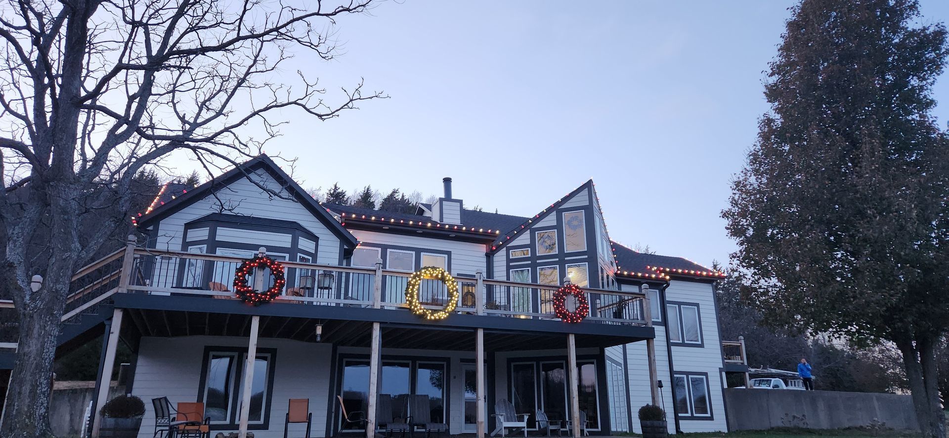 A large, white house decorated with Christmas lights and wreaths, on a deck, trees in the background.