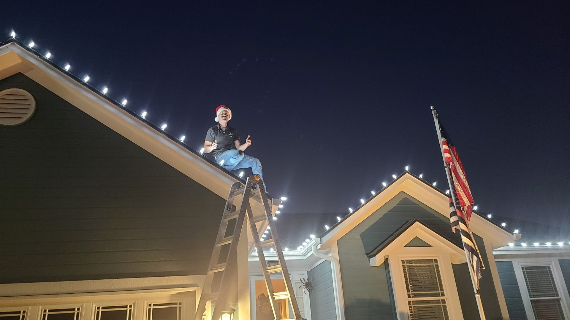 Person on ladder placing Christmas lights on a two-story house roof, night.
