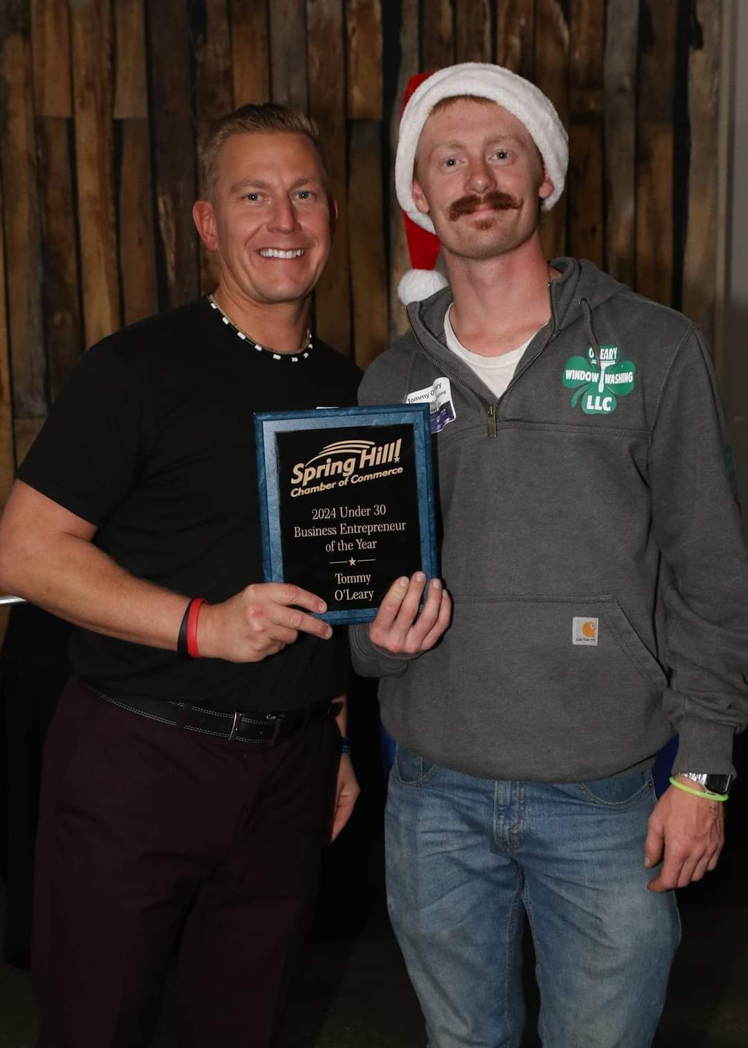 Two men holding an award; one in a Santa hat and hoodie, the other in a black t-shirt.