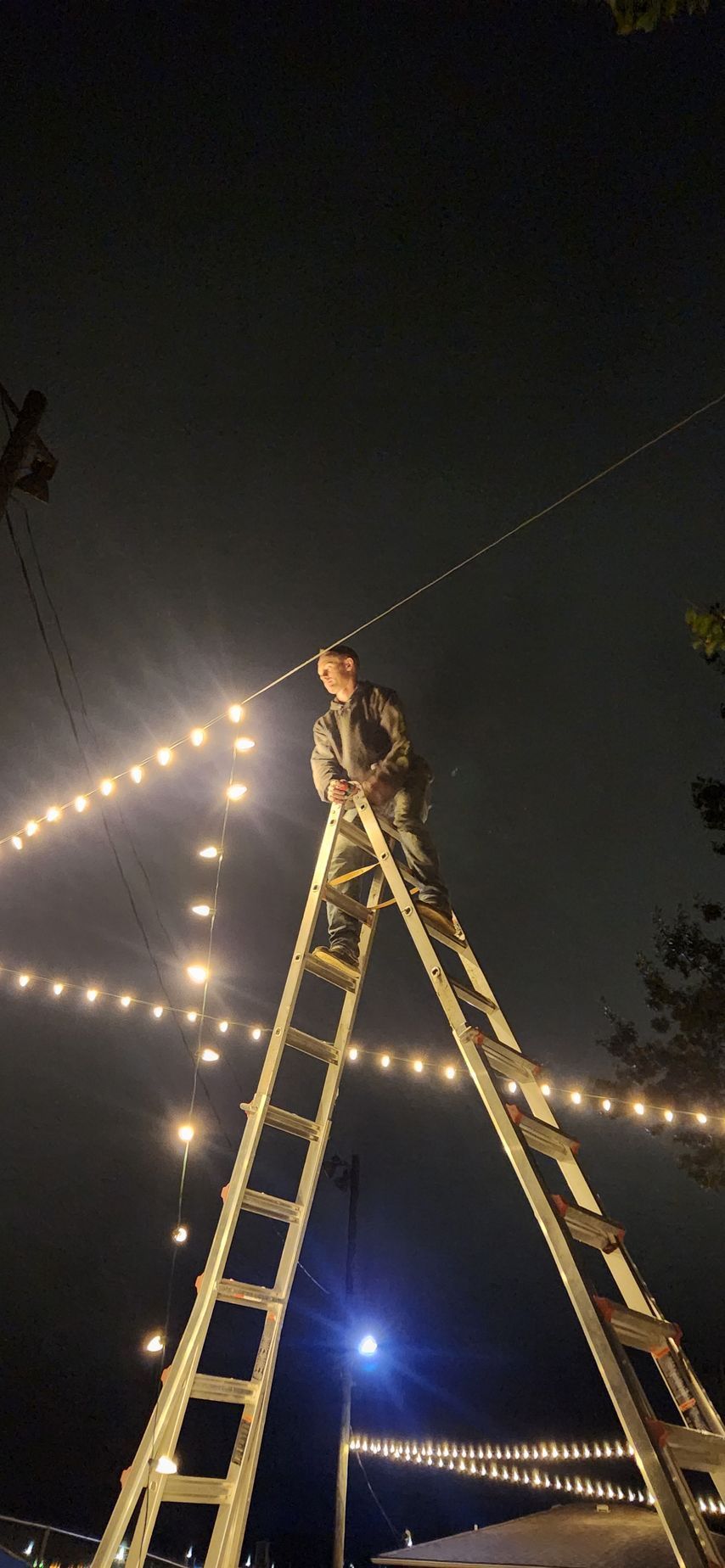 A person on a ladder hangs lights at night.