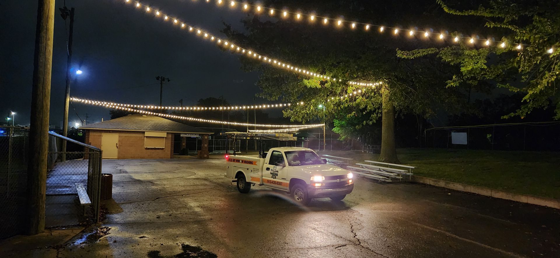 White pickup truck driving on a wet road, under strings of lights at night.