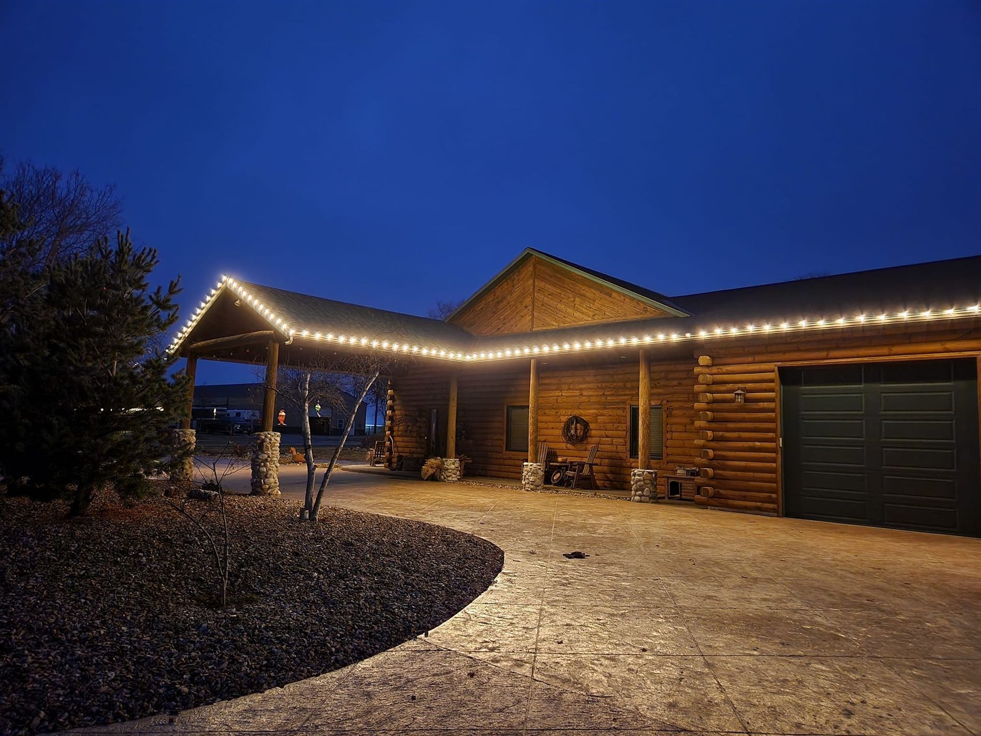 Log cabin at dusk with string lights along the roofline, gravel driveway, and dark blue sky.