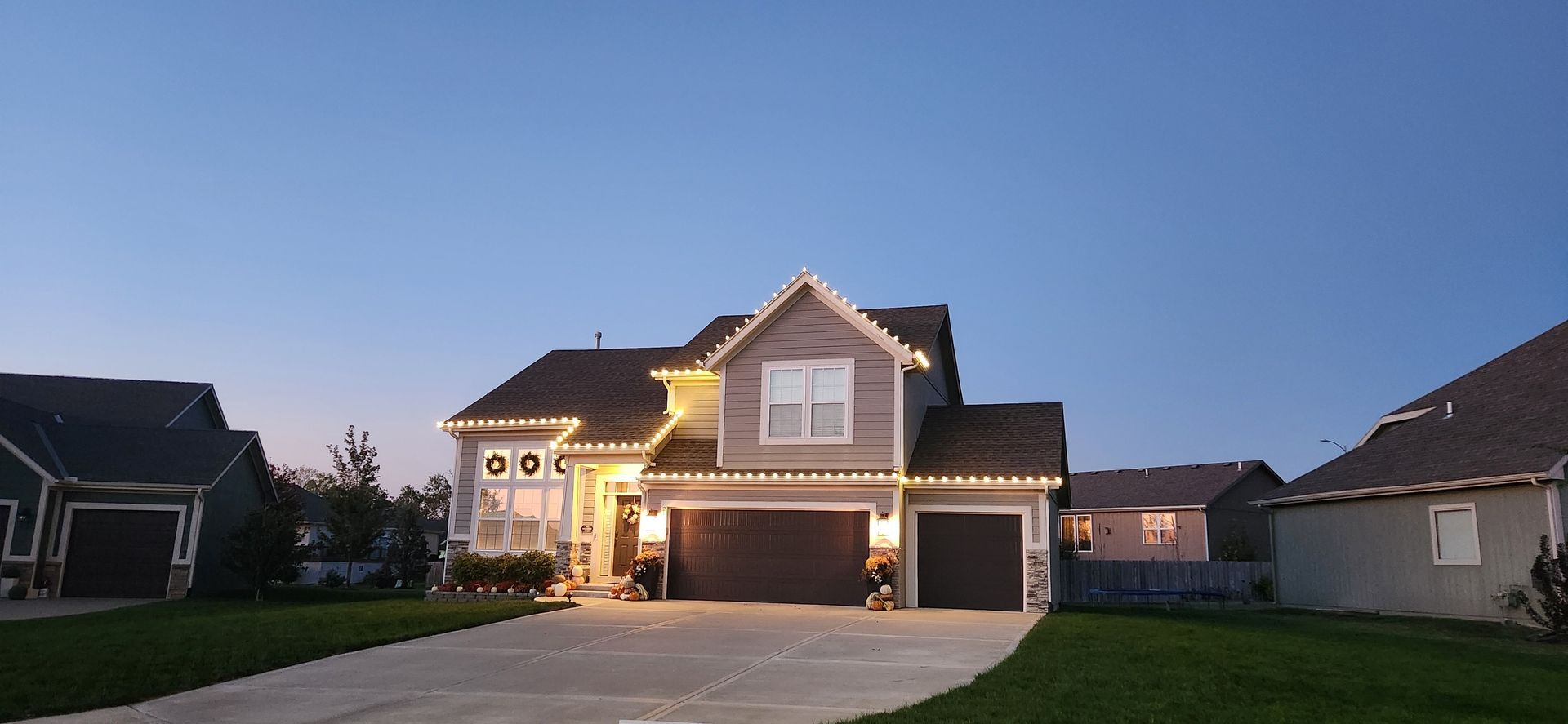 House with lights illuminated at dusk, with a driveway and lawn, in a neighborhood setting.