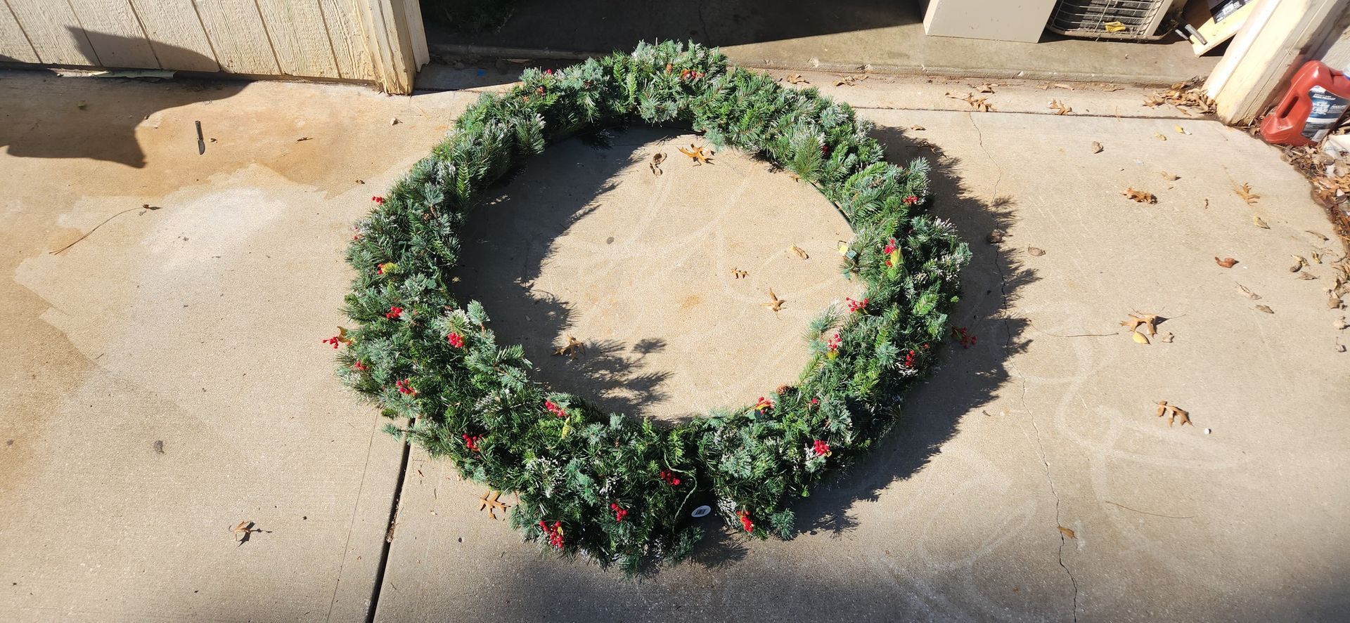 A large, circular Christmas wreath with red berries lies on a concrete surface, near a building.
