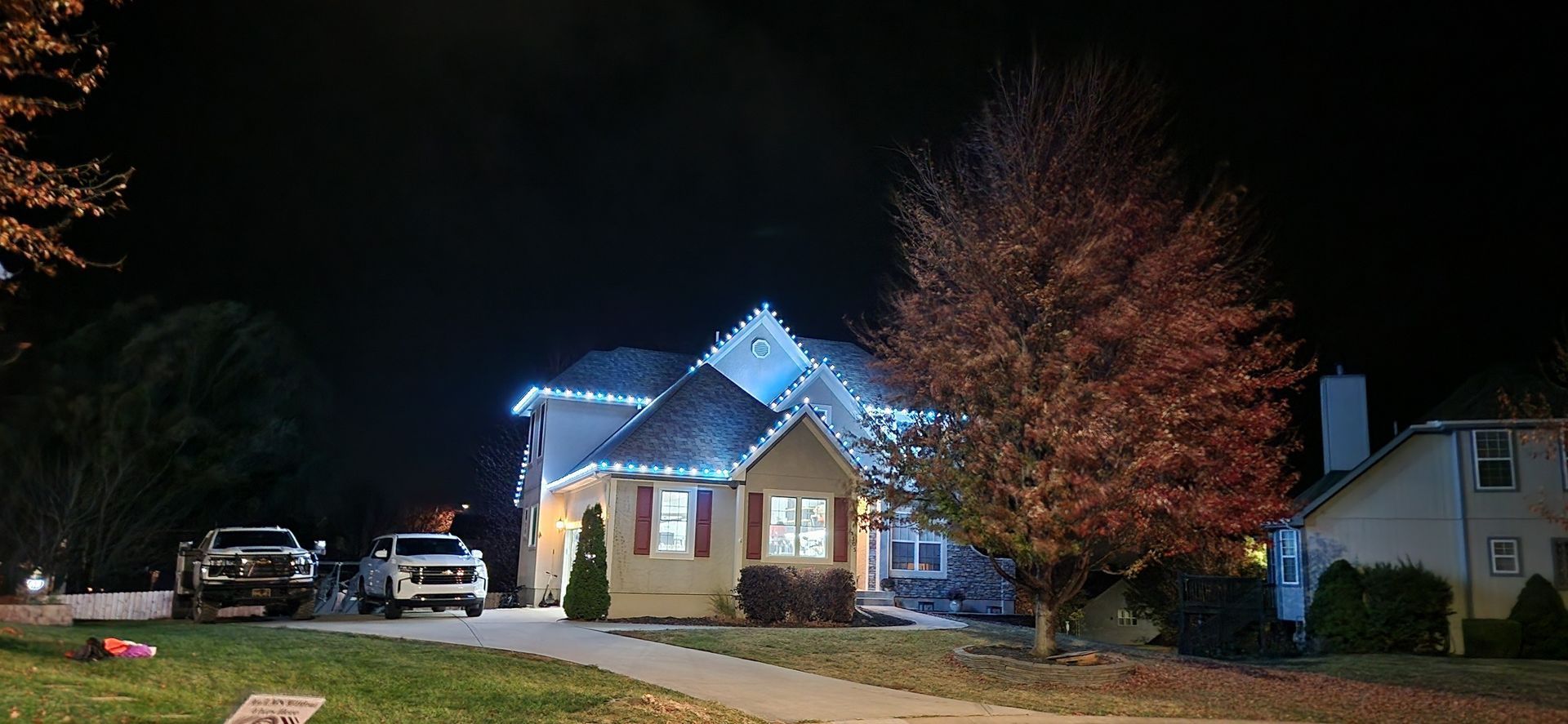 A house at night, roof and trim decorated with blue lights. Two trucks in driveway. Tree with red leaves.