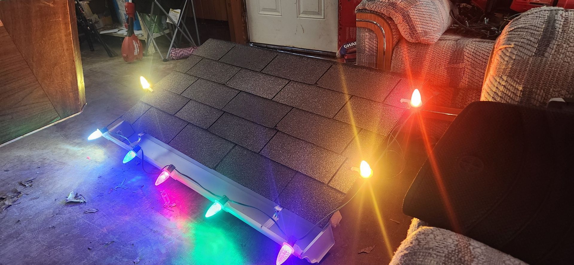 A miniature roof with colorful Christmas lights, set on a floor with soft lighting and a couch visible.
