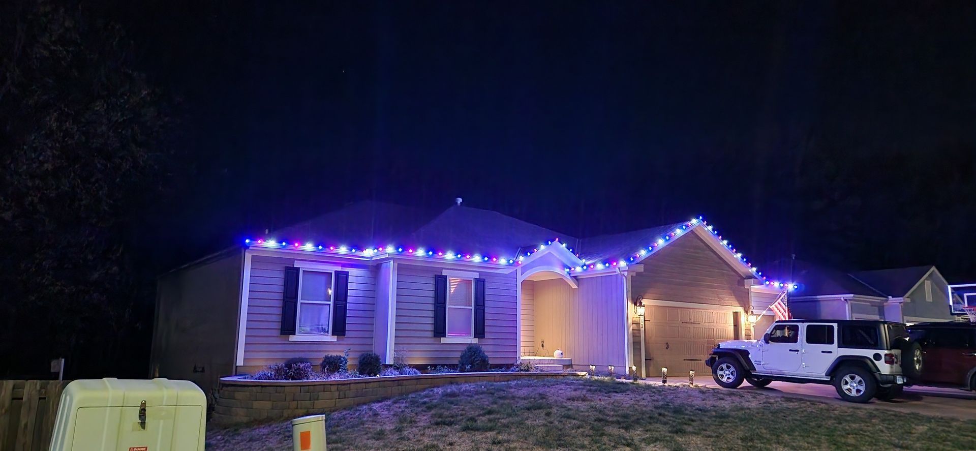 A house at night with blue Christmas lights, a white jeep in the driveway, and dark sky.
