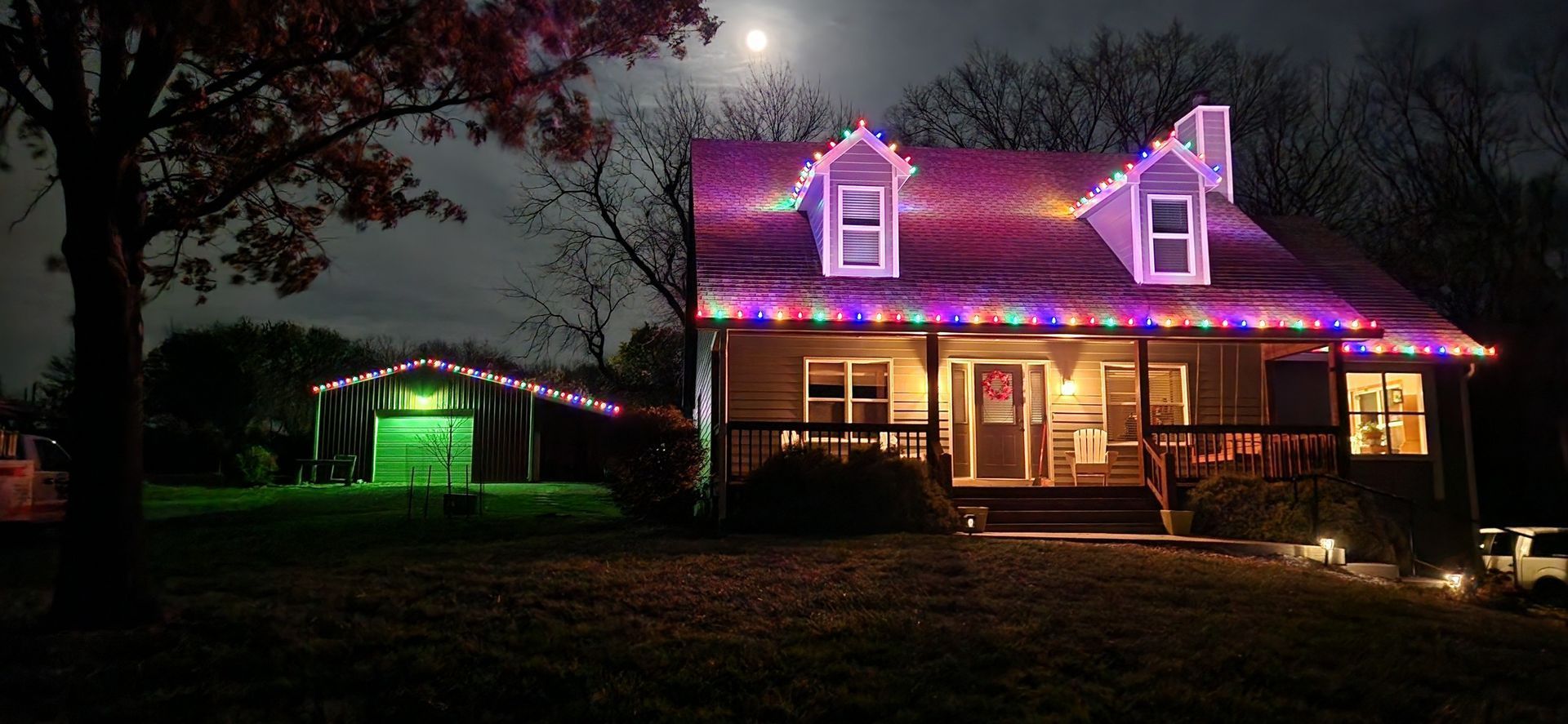 A house decorated with colorful lights at night, with a barn illuminated in green nearby.