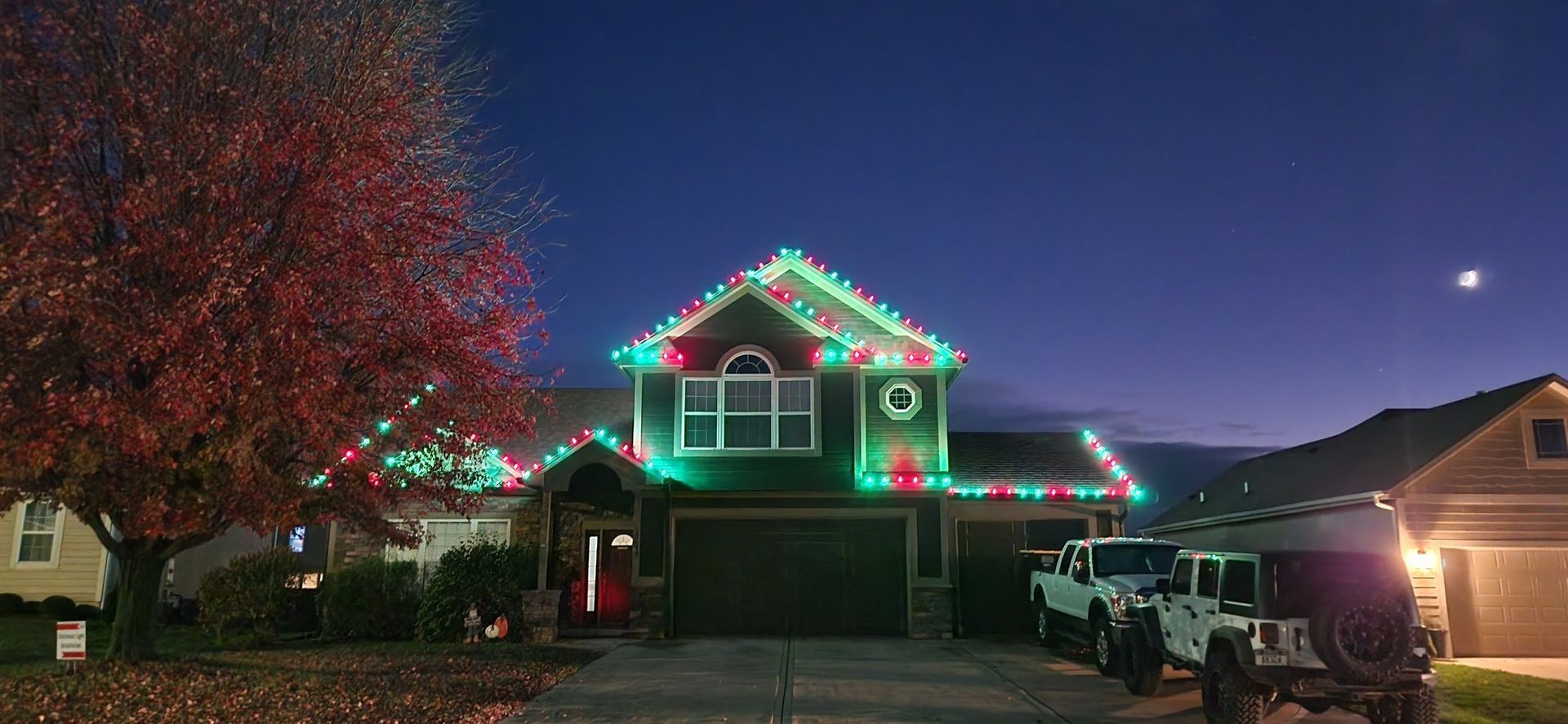 House decorated with Christmas lights at dusk, lit tree in the foreground. Two jeeps parked in the driveway.