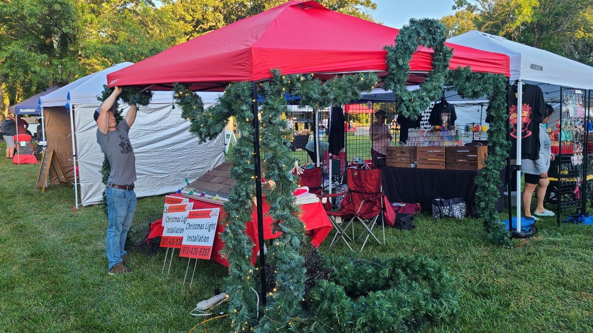 Man decorates a red tent at an outdoor market with green garland. Other booths and shoppers are visible.