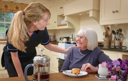 An elderly woman is sitting at a table with a plate of food and a nurse is standing next to her.