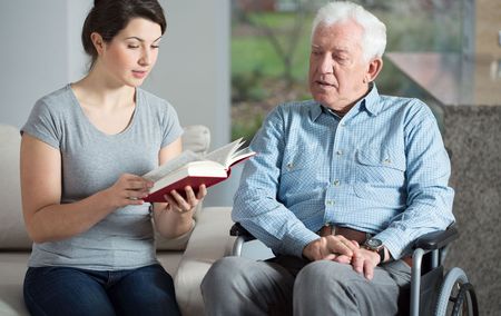 A woman is reading a book to an elderly man in a wheelchair.