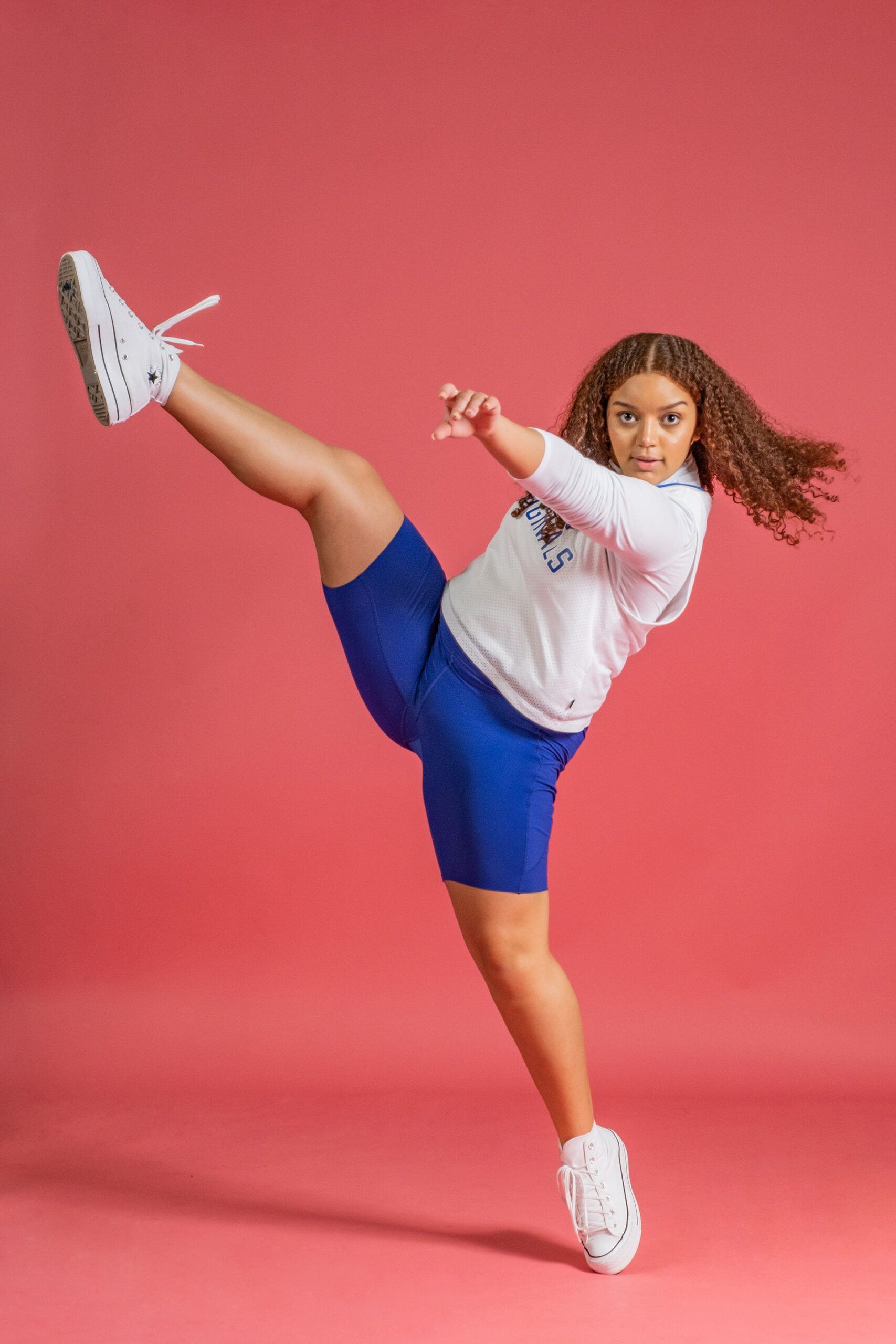 A young girl is standing on one leg in front of a pink background.