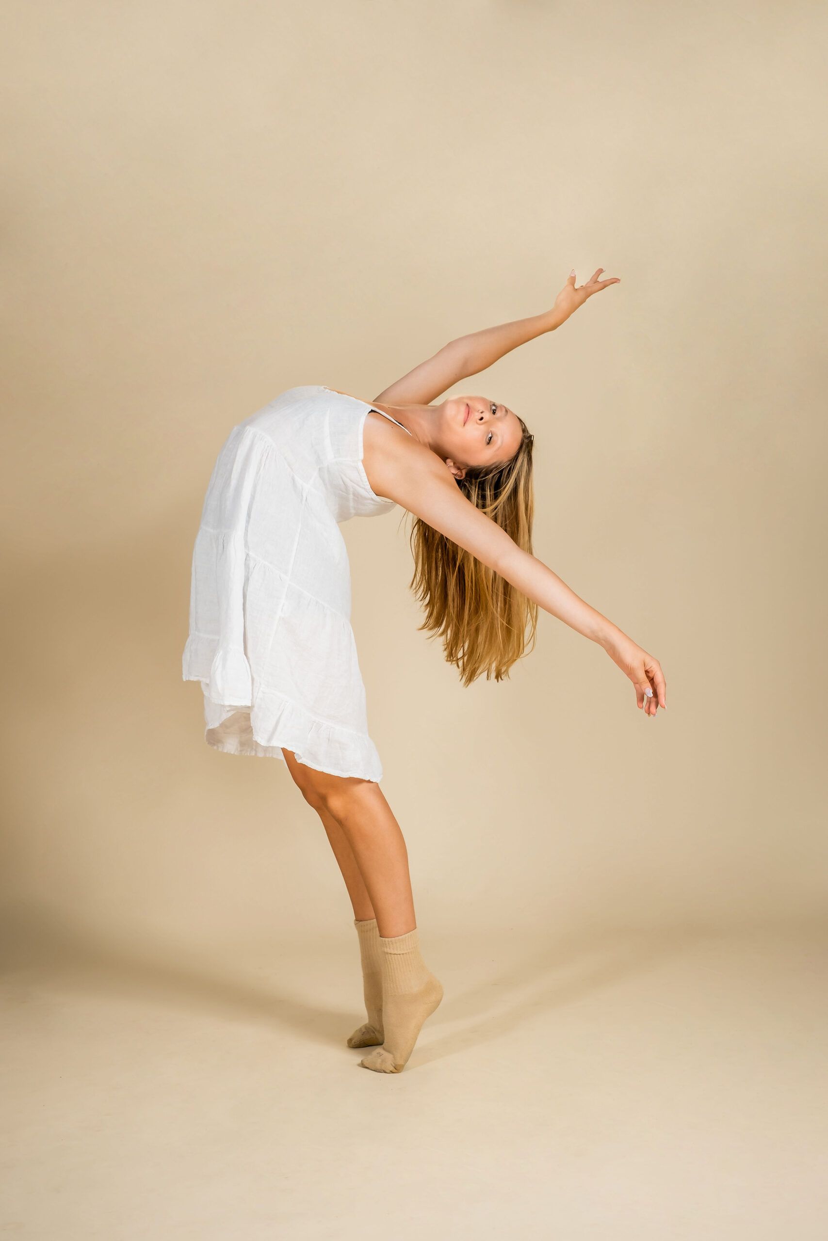 A young woman in a white dress is dancing on a beige background.