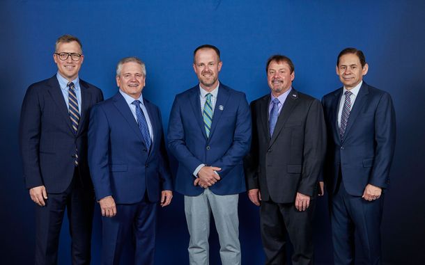Five men in suits pose standing together against a blue studio backdrop.