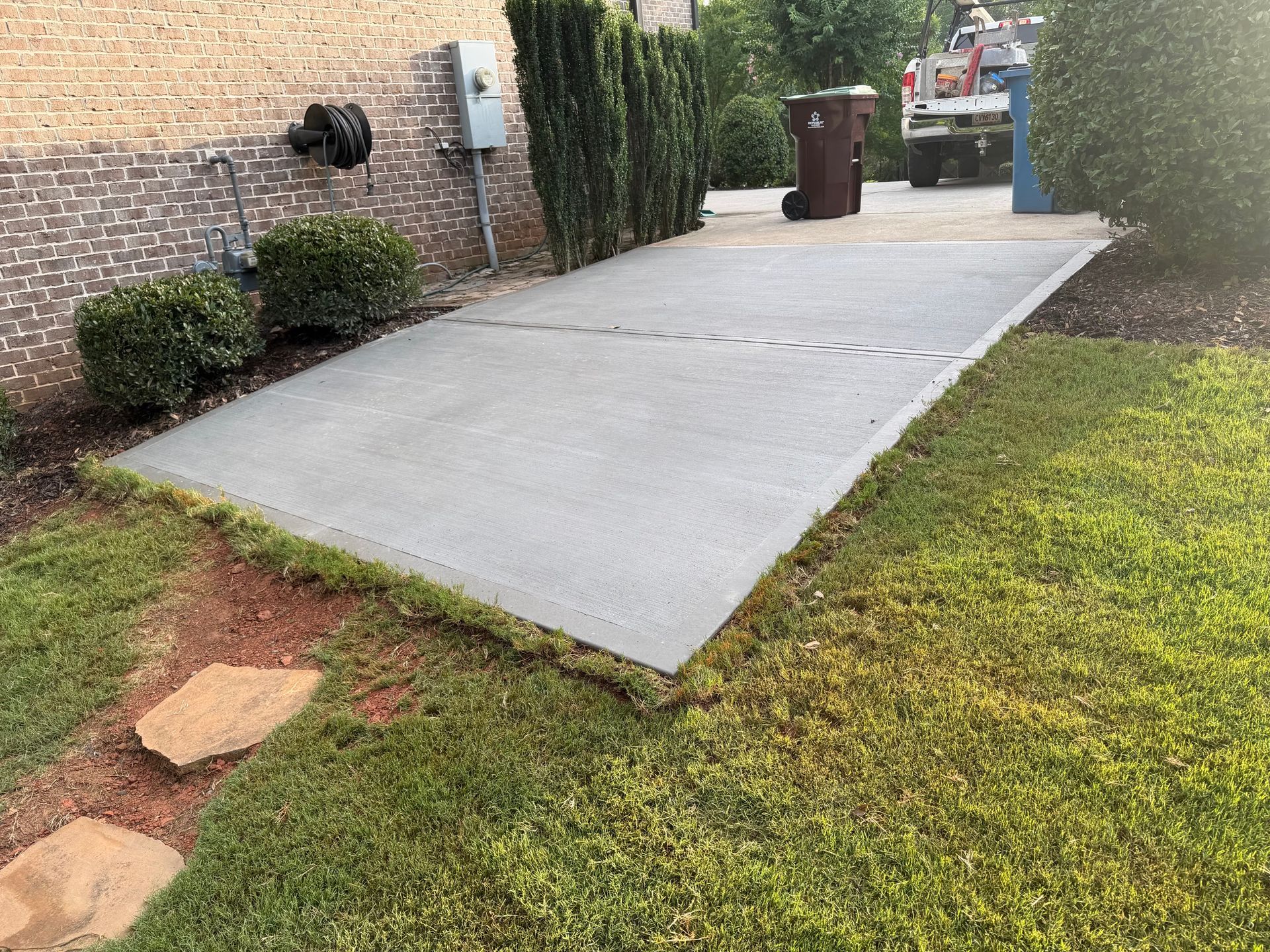 Concrete driveway beside a brick house, bordered by grass and shrubs, leading to a trash bin at the back.