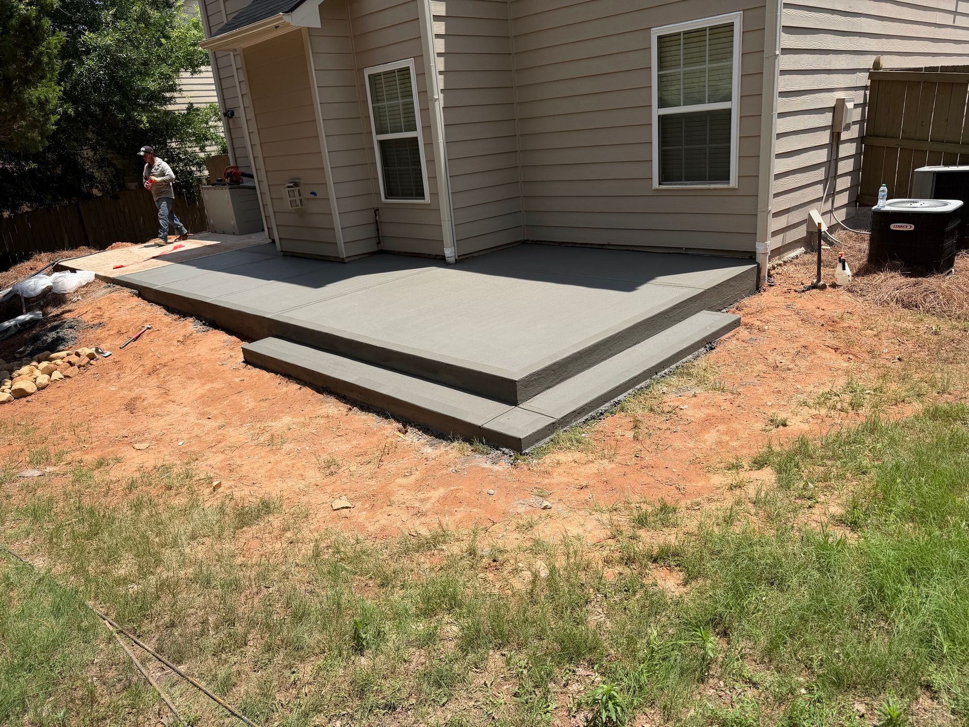 Backyard concrete patio with steps beside a house, bordered by dirt and grass.