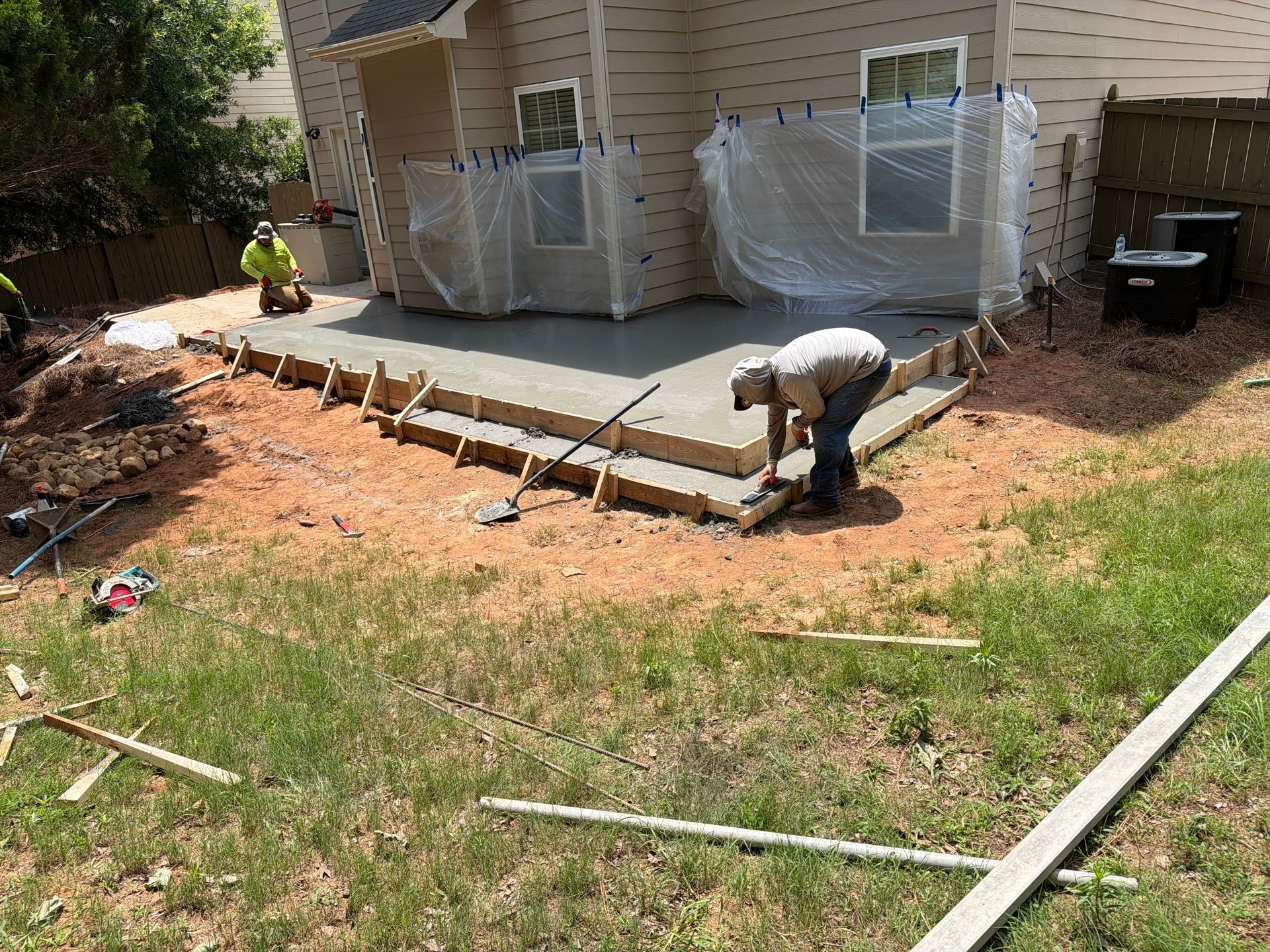 Worker building a concrete patio ramp beside a beige house, with windows wrapped in plastic.