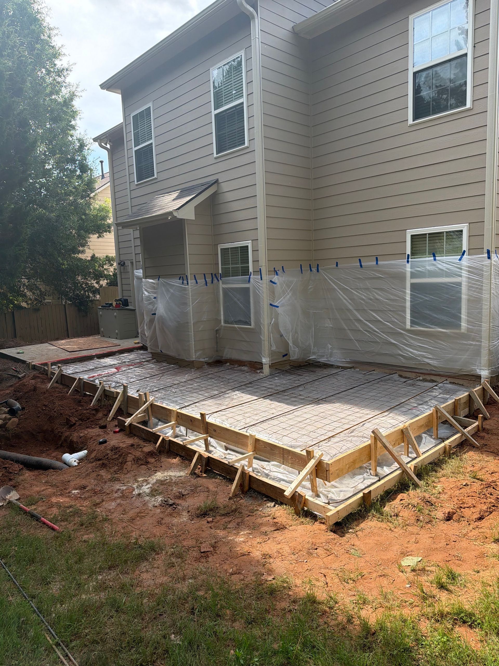 Concrete patio under construction beside a beige two-story house, with wooden forms and exposed soil around it