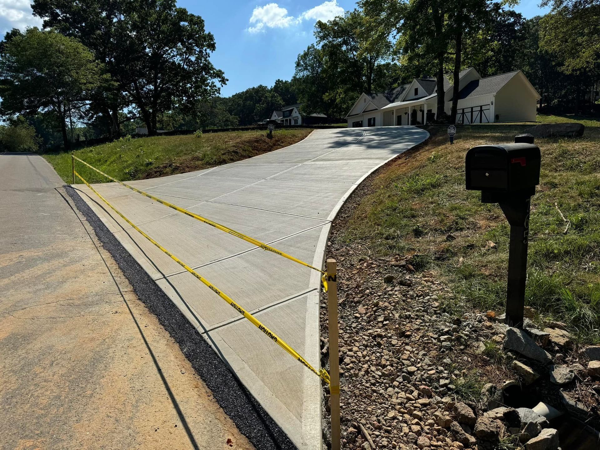 Newly paved driveway with yellow caution tape, leading to a white house under a sunny sky.