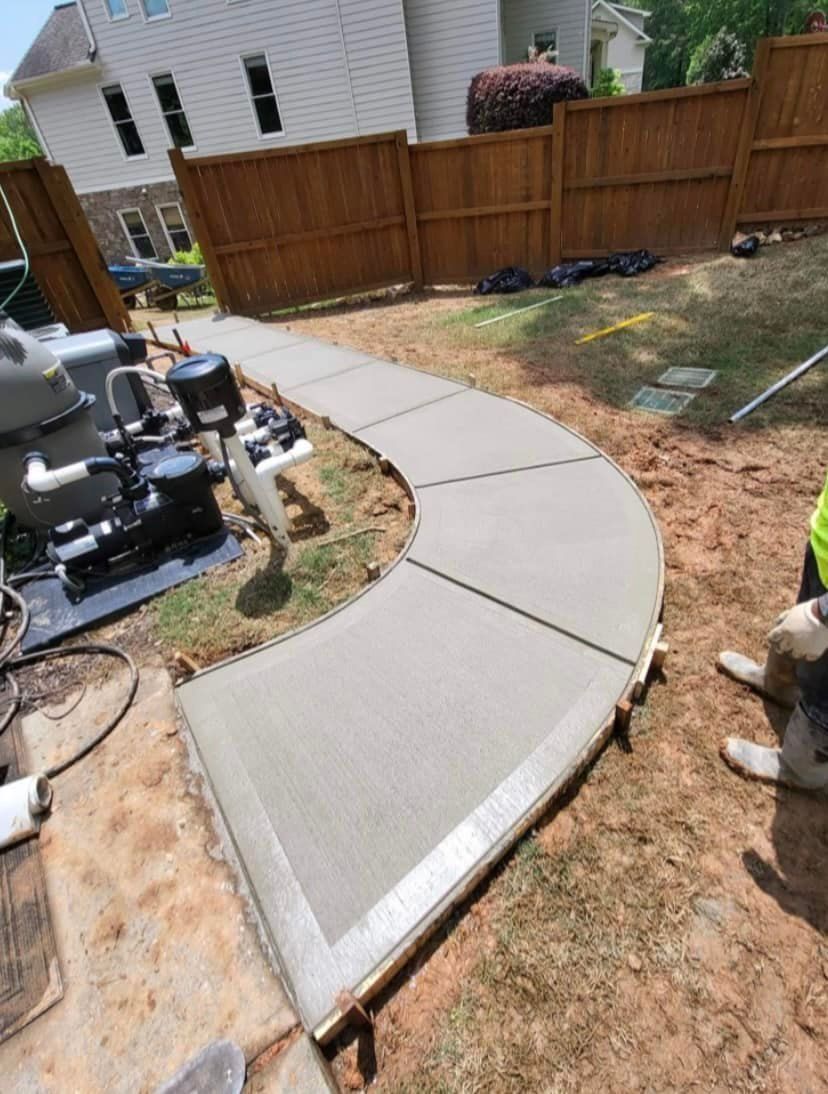 Curving concrete walkway under construction near a fence and pool equipment. Light gray and brown hues.