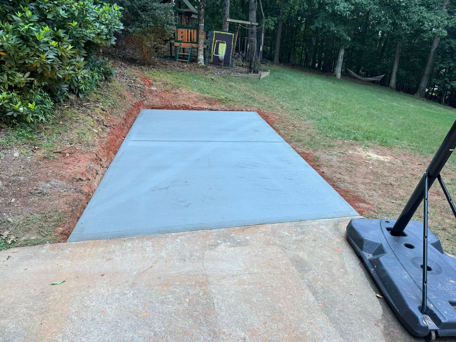 A rectangular concrete pad in a yard, bordered by dirt. A basketball hoop is visible.