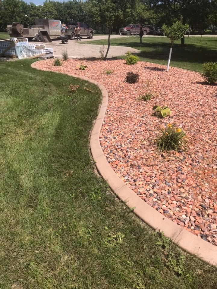 A landscaped garden bed with red gravel and green plants, bordered by a concrete curb, next to green grass.
