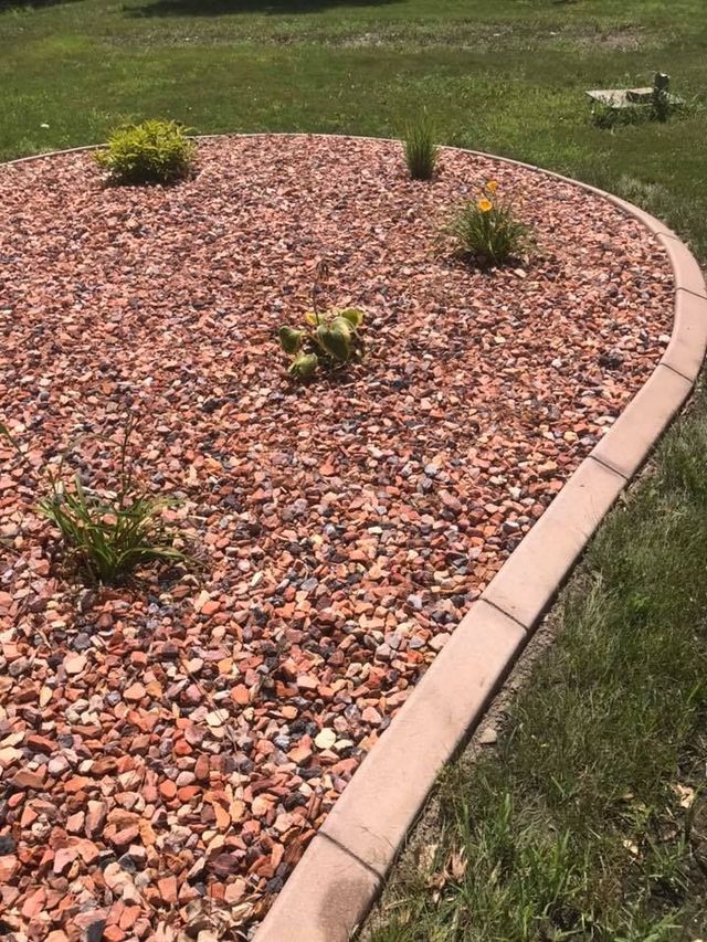 Curved flower bed with reddish gravel, concrete edging, and green plants. Lawn surrounds the bed.