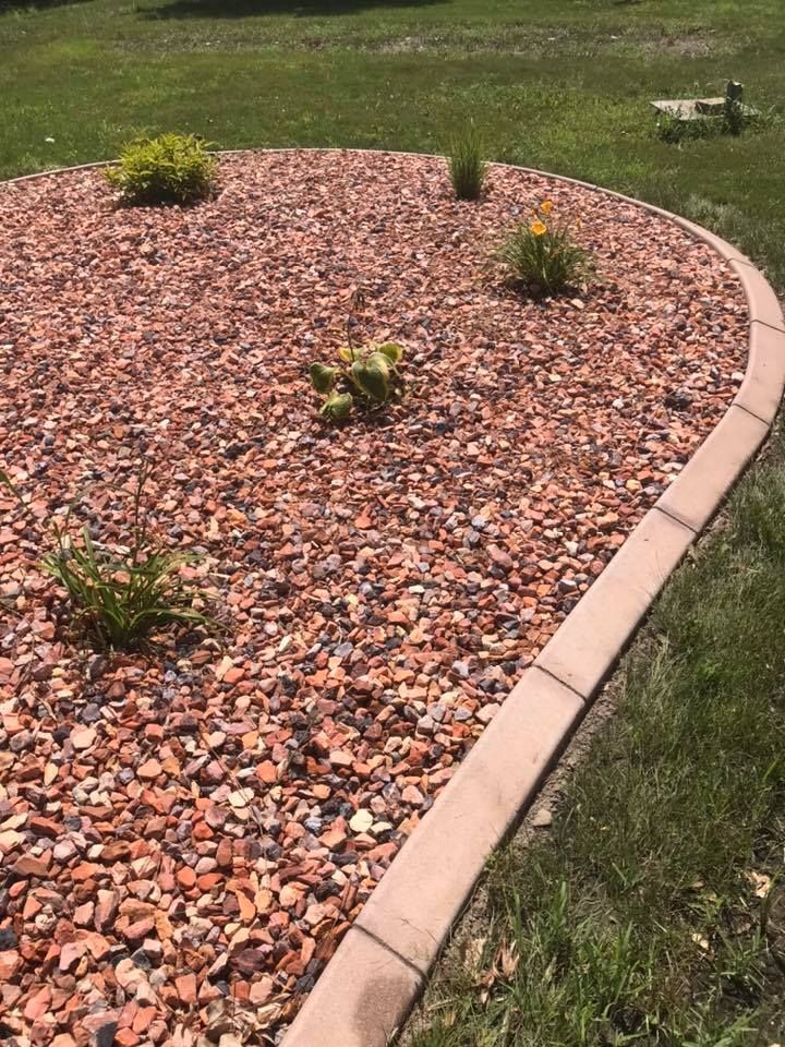 Curved garden bed filled with red gravel, bordered by concrete, and containing a few small plants.
