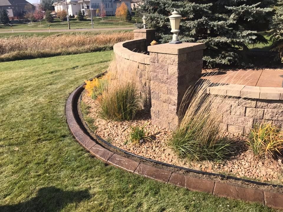 Curved retaining wall with grass, gravel, and plants near a lawn and pond.