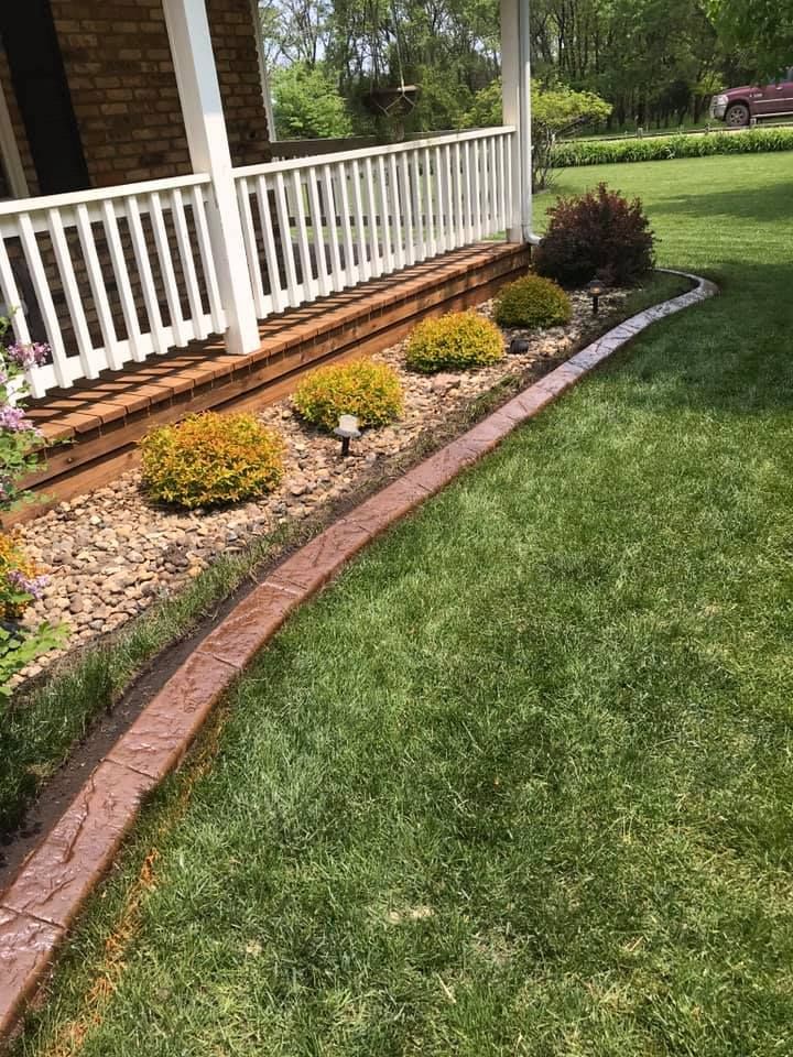Brown concrete curbing borders a flower bed of green and golden shrubs, next to green lawn, white porch, and brick house.