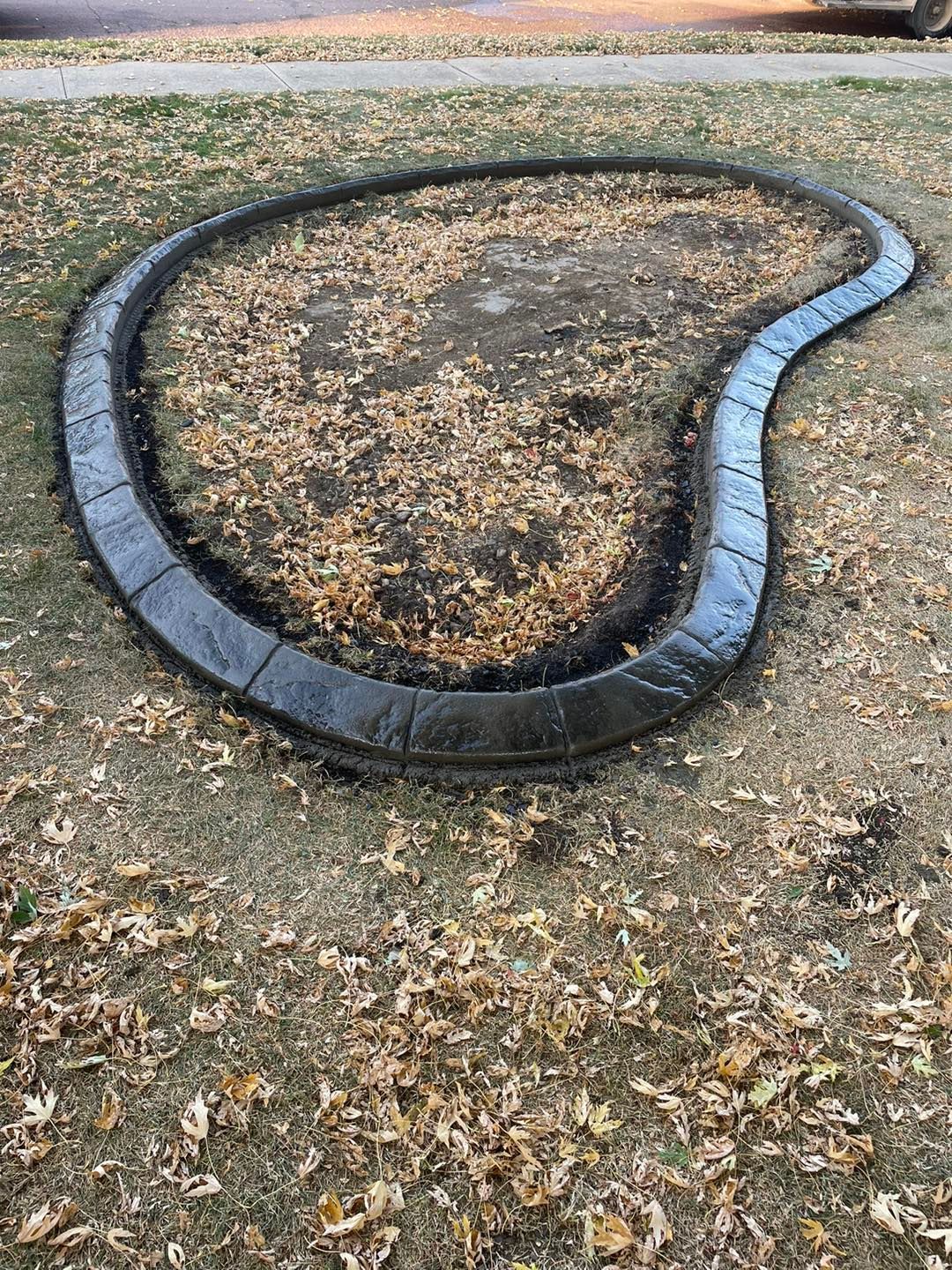 Dark gray concrete curbing outlines a leaf-covered garden bed on a lawn.