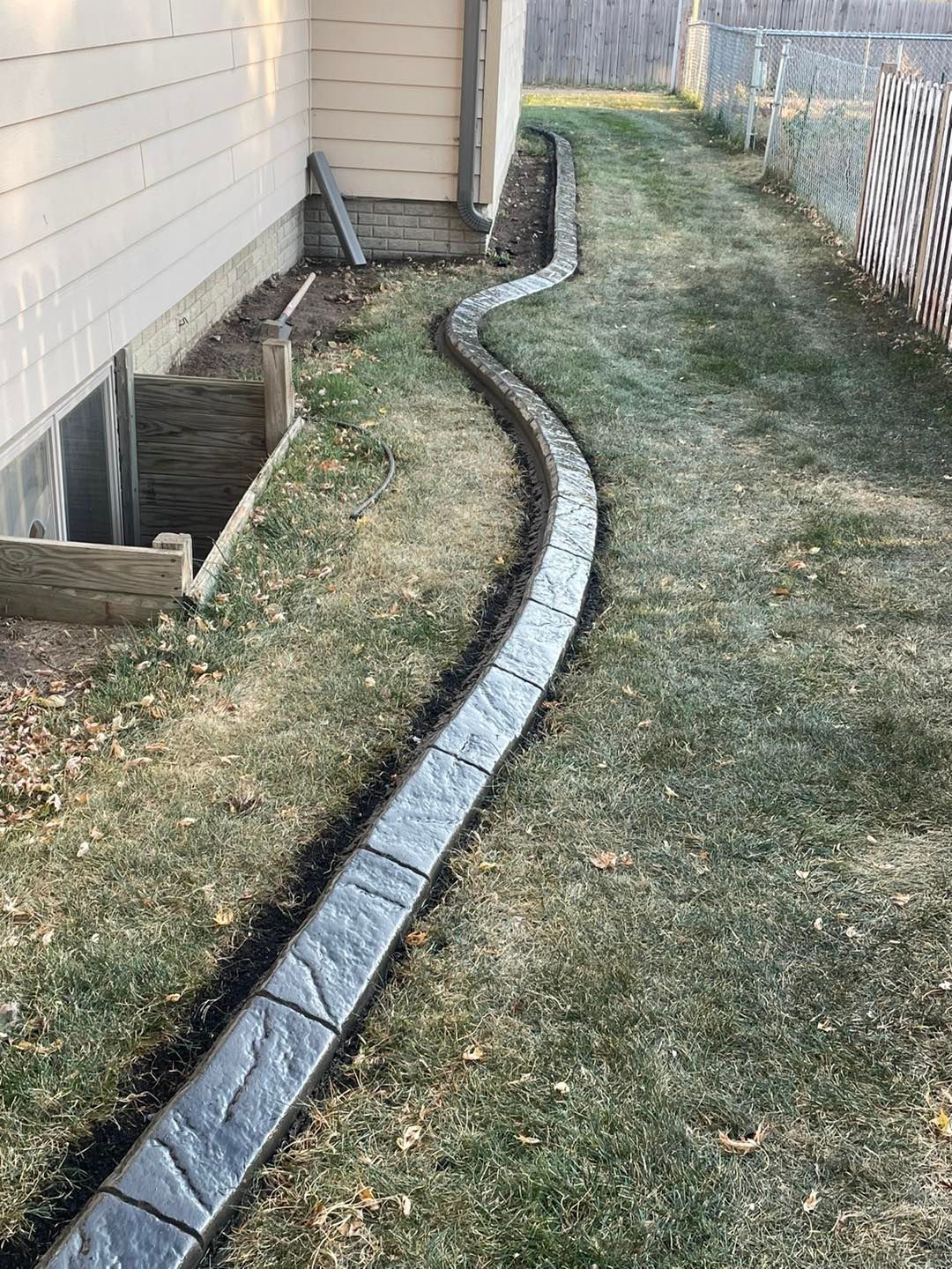 Curved, textured concrete border defines a garden bed, filled with fallen leaves, in a grassy yard.