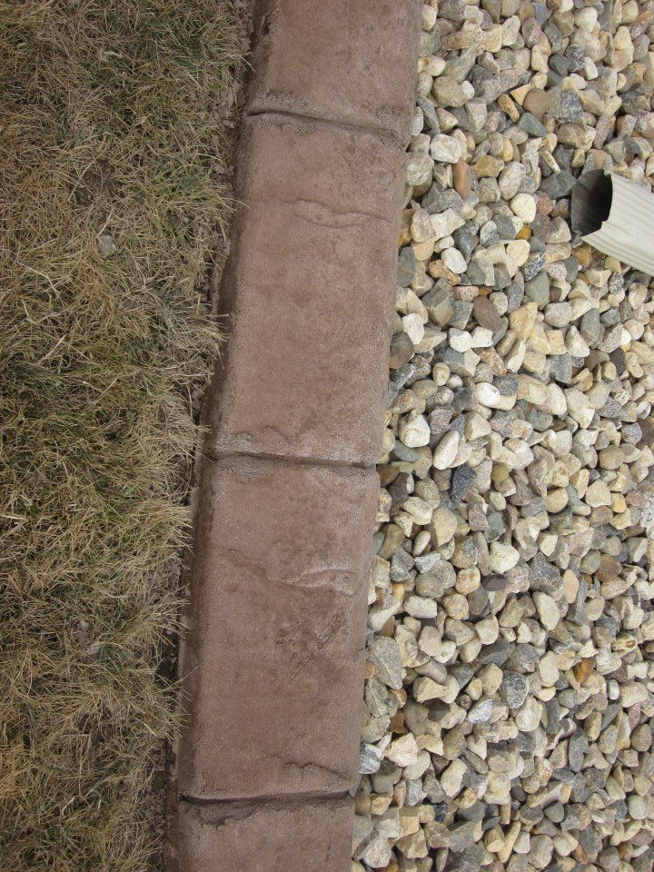 Brown concrete edging separates dry grass and gravel. A downspout is visible on the right.