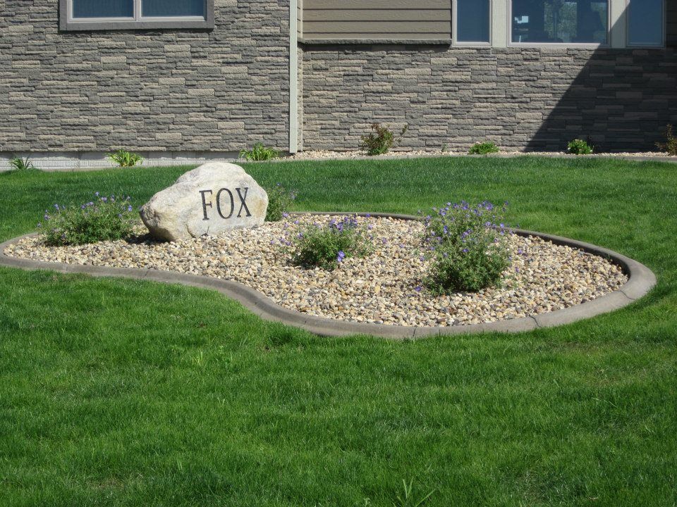 Rock with FOX in a beige gravel bed, surrounded by a concrete border and grass, in front of a house.
