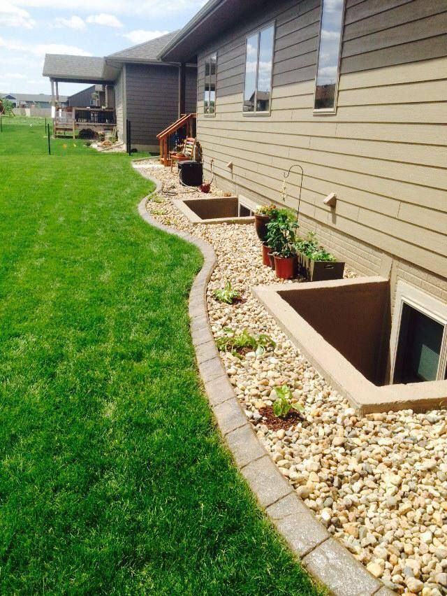 Backyard with house and a decorative stone border separating lawn and window wells filled with gravel and potted plants.