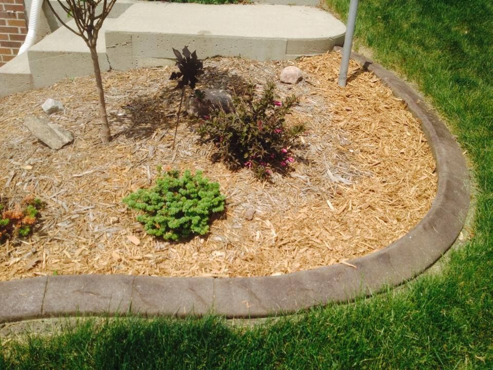 Brown mulch bed with plants, bordered by gray concrete curbing, beside green grass.