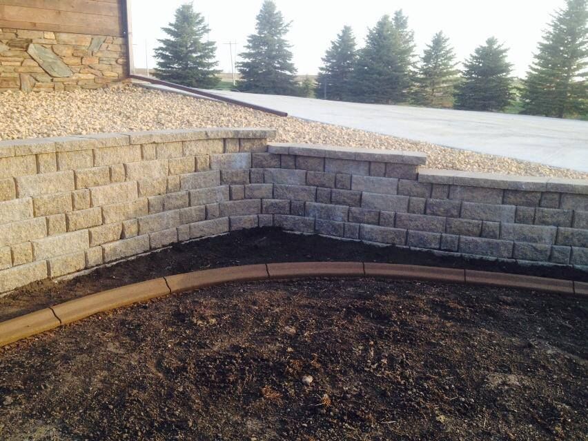 Stone retaining wall with curved brown border, gravel, and dark soil. Evergreen trees in the background.