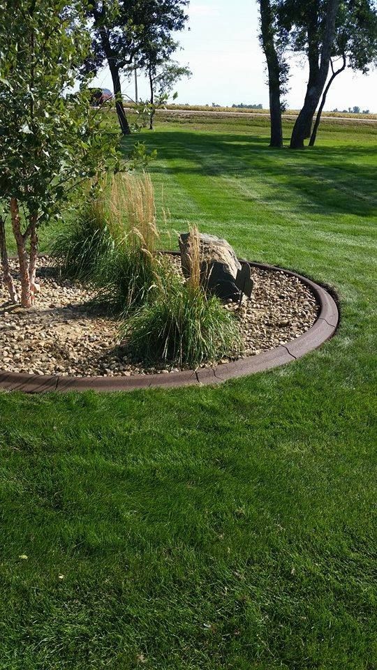 Lawn with a bed of rocks and plants, edged with brown border. Green grass, trees in the background.