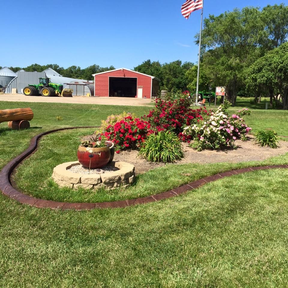 A red-roofed barn and a green John Deere tractor are in the background of a floral garden with a small fountain.