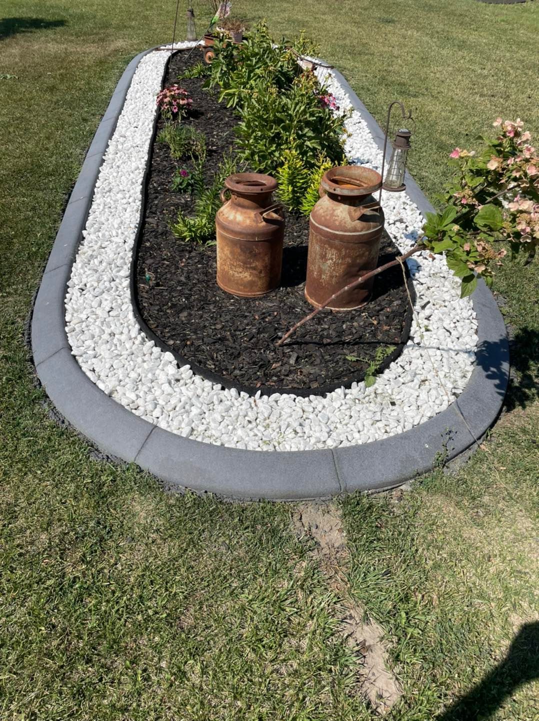 A curved garden bed with black mulch, white stones, and rusted metal milk cans, framed by a gray border, on a grassy lawn.